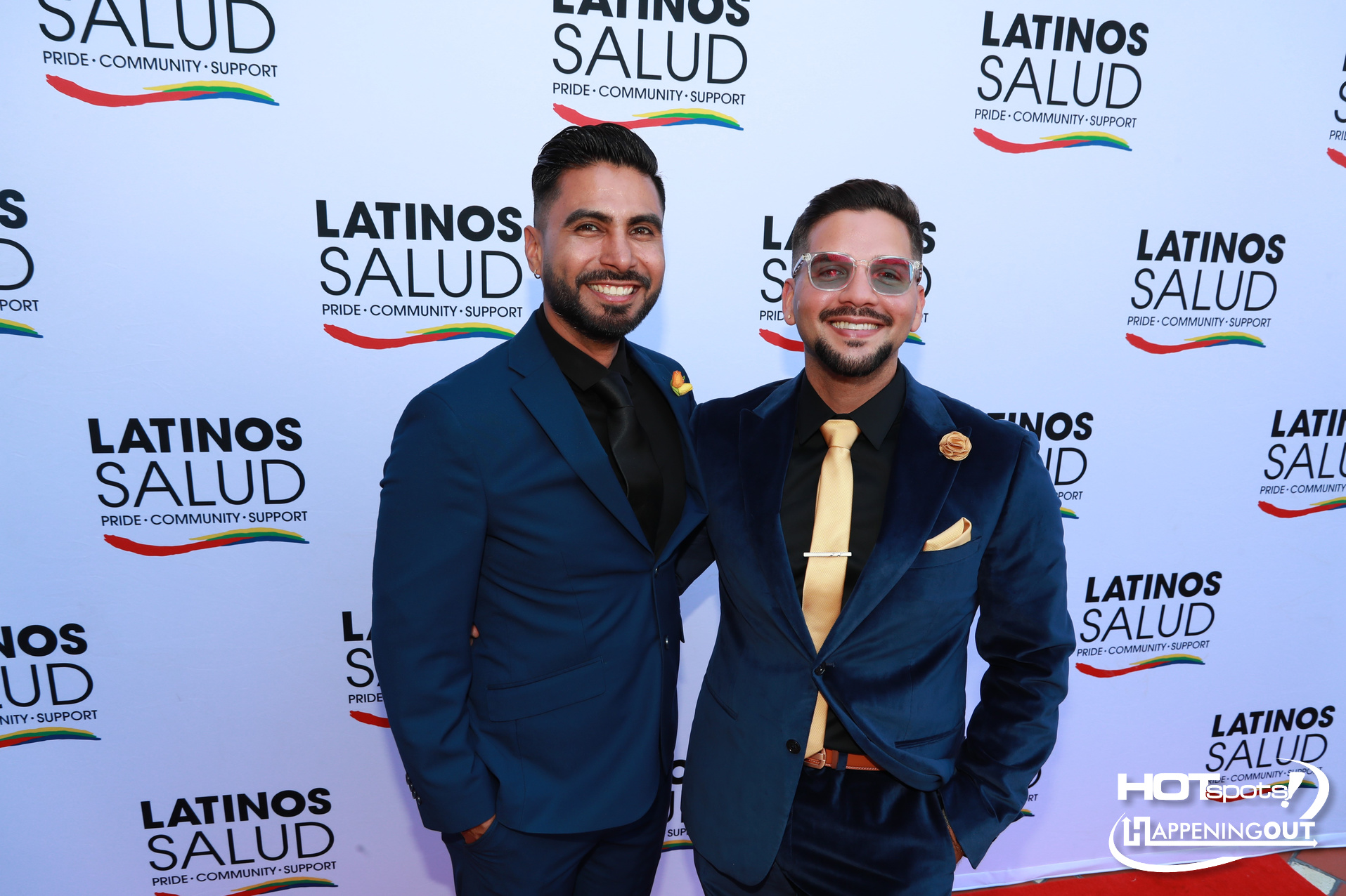 Two men in blue suits posing on a red carpet at a Latinos Salud event, backdrop repeats the organization’s logo.