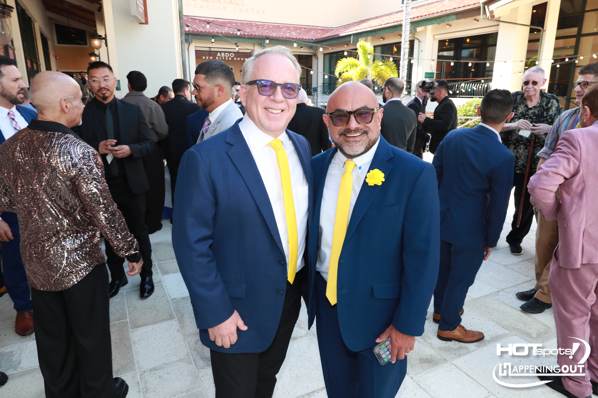 Two men in blue suits with yellow ties smile for a photo at an outdoor social event, with guests mingling behind them and event logos in the corner.
