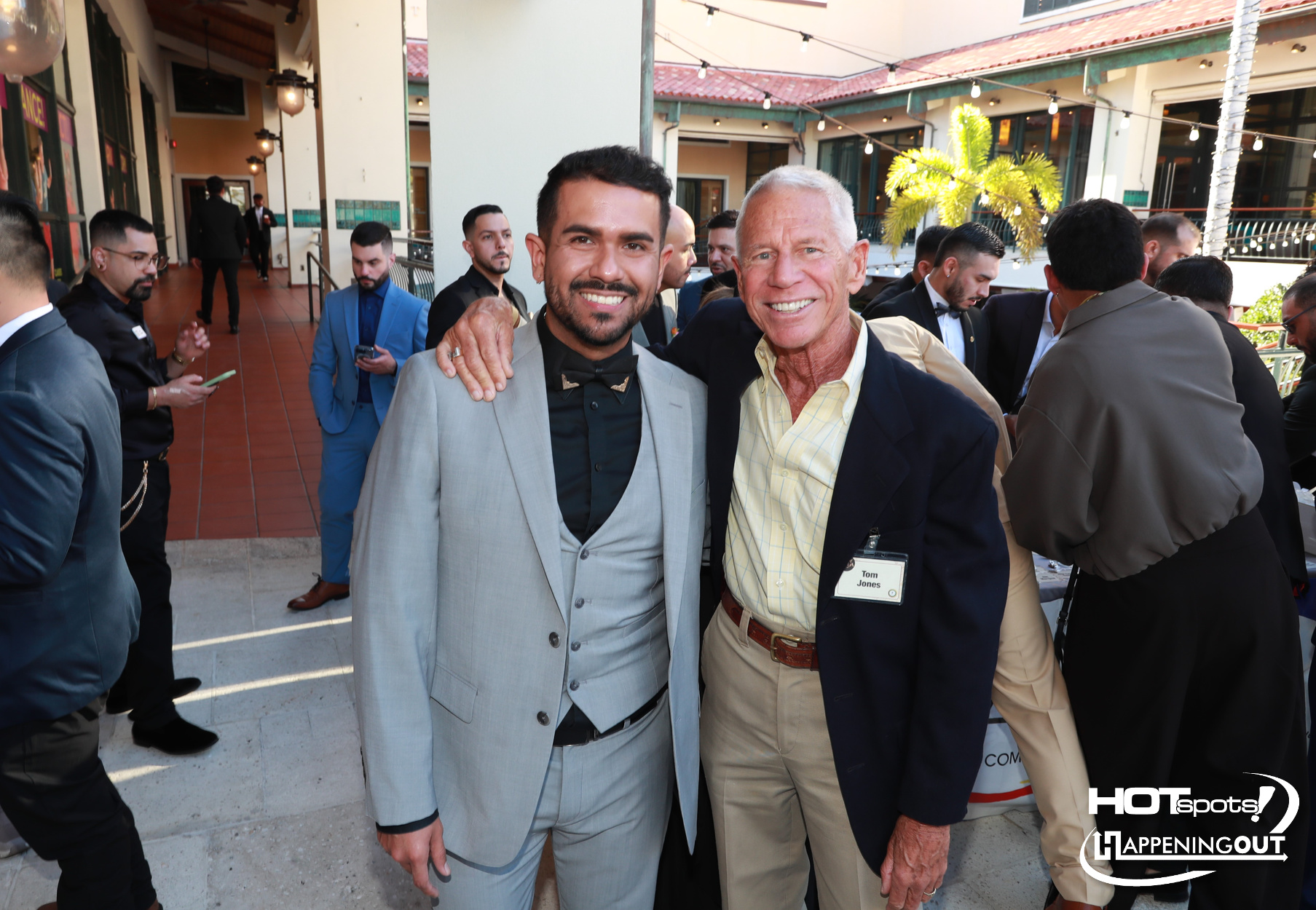 Two men smile for a photo at a formal outdoor event, one in a light gray suit with a bow tie and the other in a dark blazer with a name badge visible.