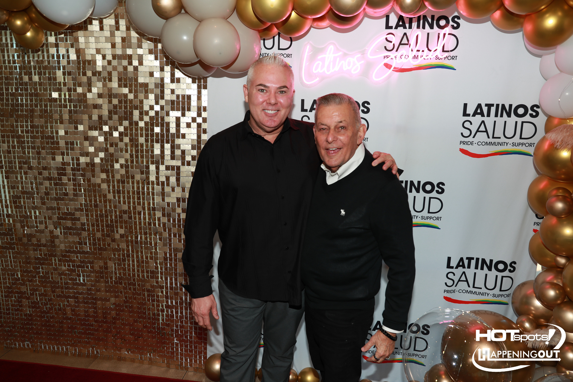 Two men pose arm-in-arm at a Latino Salud event, in front of a backdrop with balloons and a gold mosaic wall.