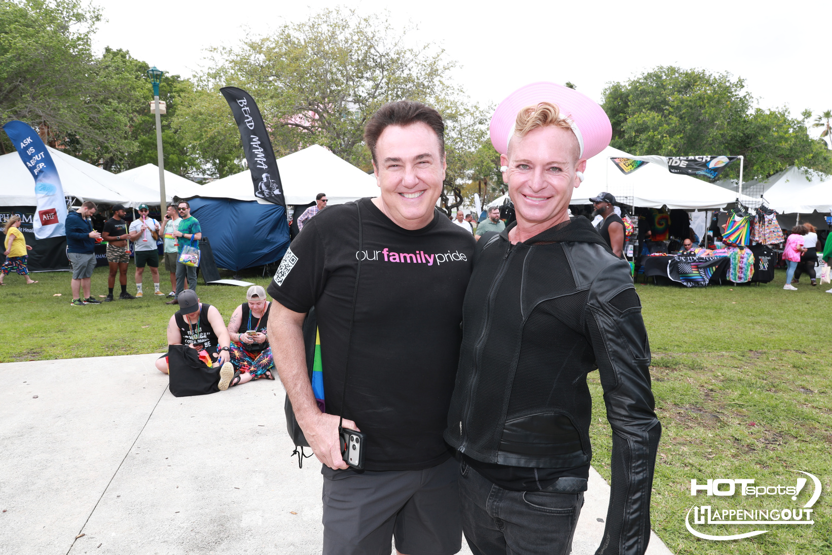 Two men pose for a photo at an outdoor festival with tents and spectators in the background, one wearing a black shirt that reads 'our family pride'.](**note:** stray char inserted)