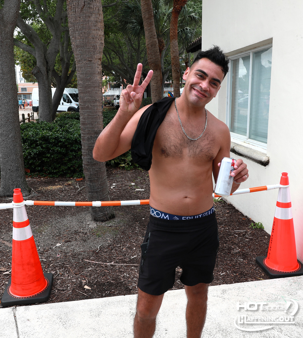 Smiling shirtless man making a peace sign, holding a can, outdoors beside orange traffic cones.