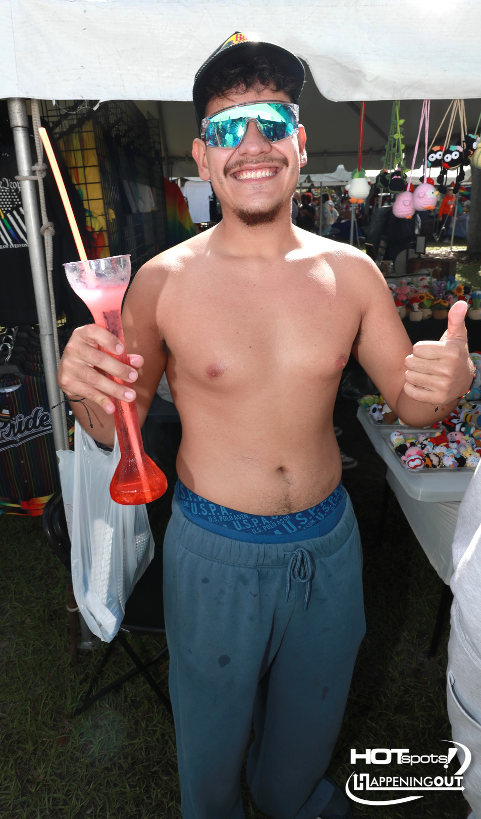 Smiling shirtless man in a cap and mirrored sunglasses holds a pink cocktail with a straw and thumbs up at an outdoor market.
