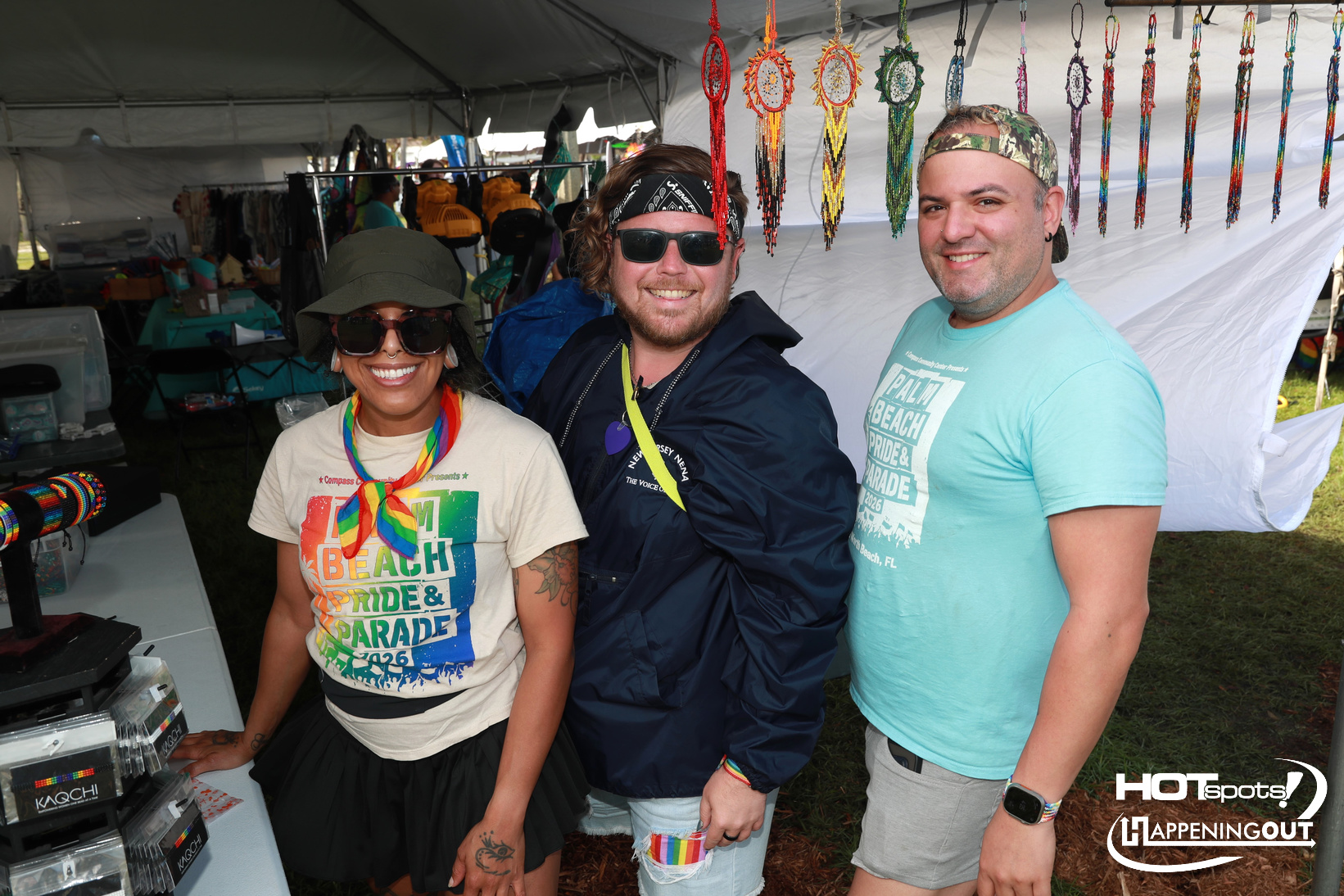 Three smiling adults stand under a tent at a Pride festival booth displaying colorful bead necklaces and accessories. A rainbow-themed scarf is worn by the person on the left, and two men in casual attire stand to the right.