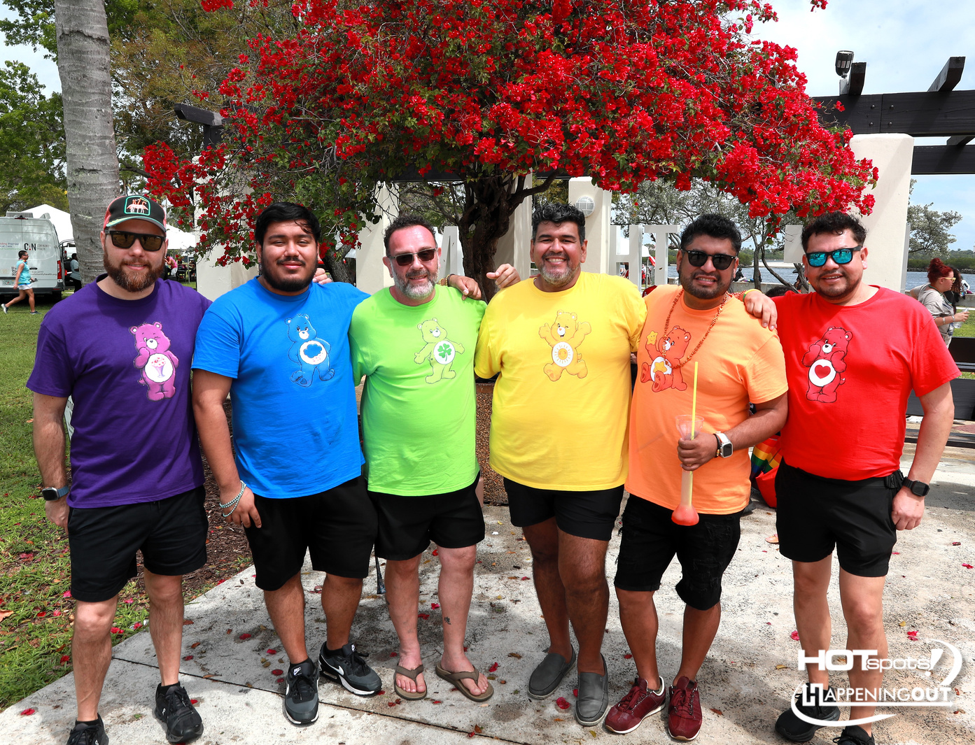 Six men in rainbow-colored T-shirts with bear logos pose together outdoors under a red flowering tree.