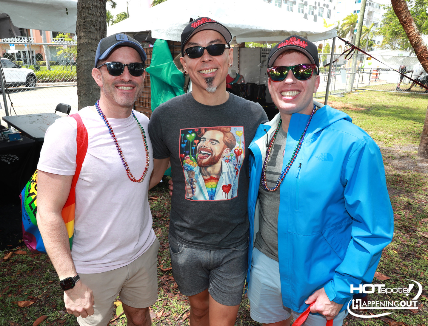 Three men posing for a photo at an outdoor event, wearing sunglasses and beaded necklaces.