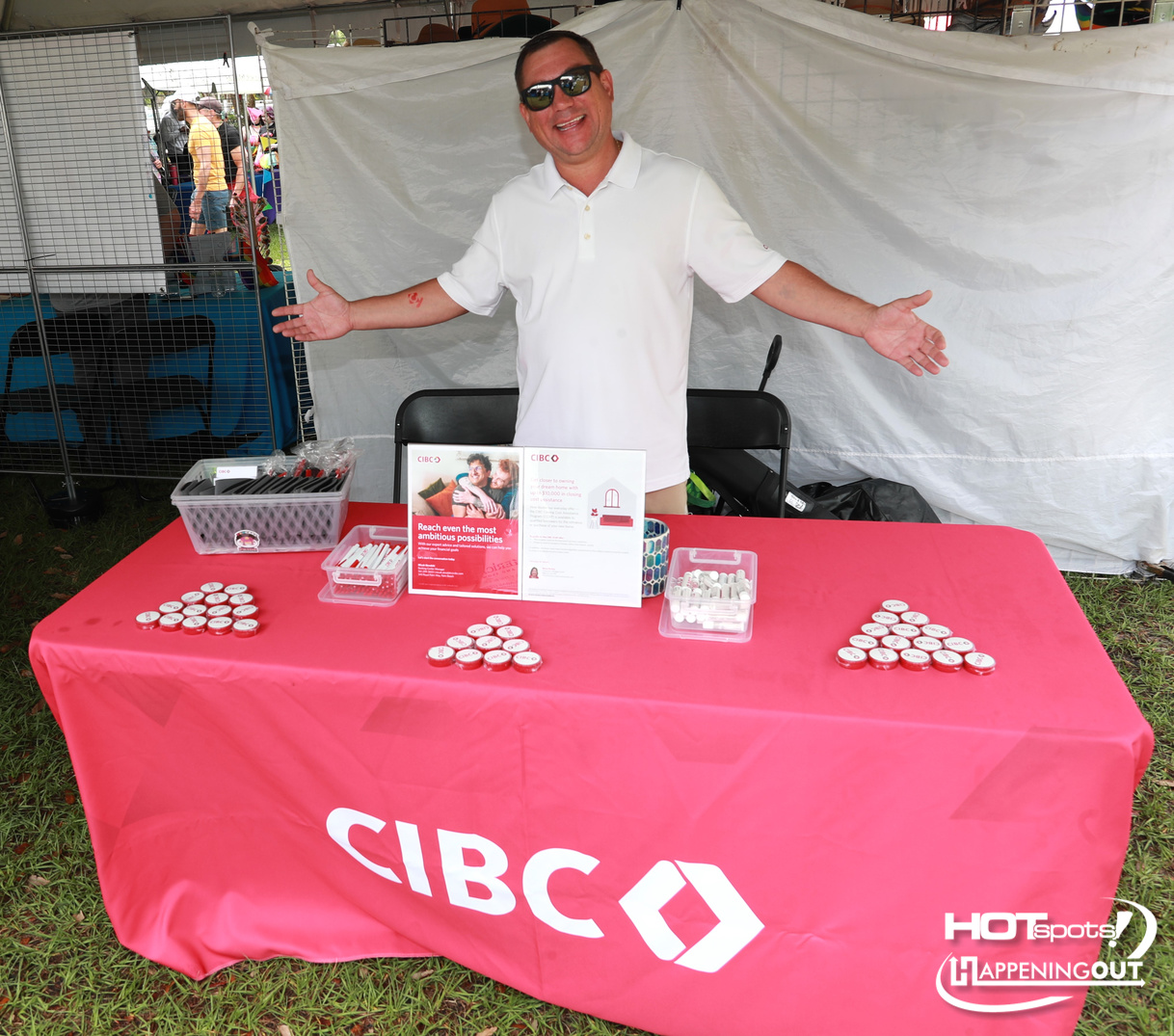 Man in white polo and sunglasses stands behind a bright pink CIBC table at an outdoor event, arms open wide.
