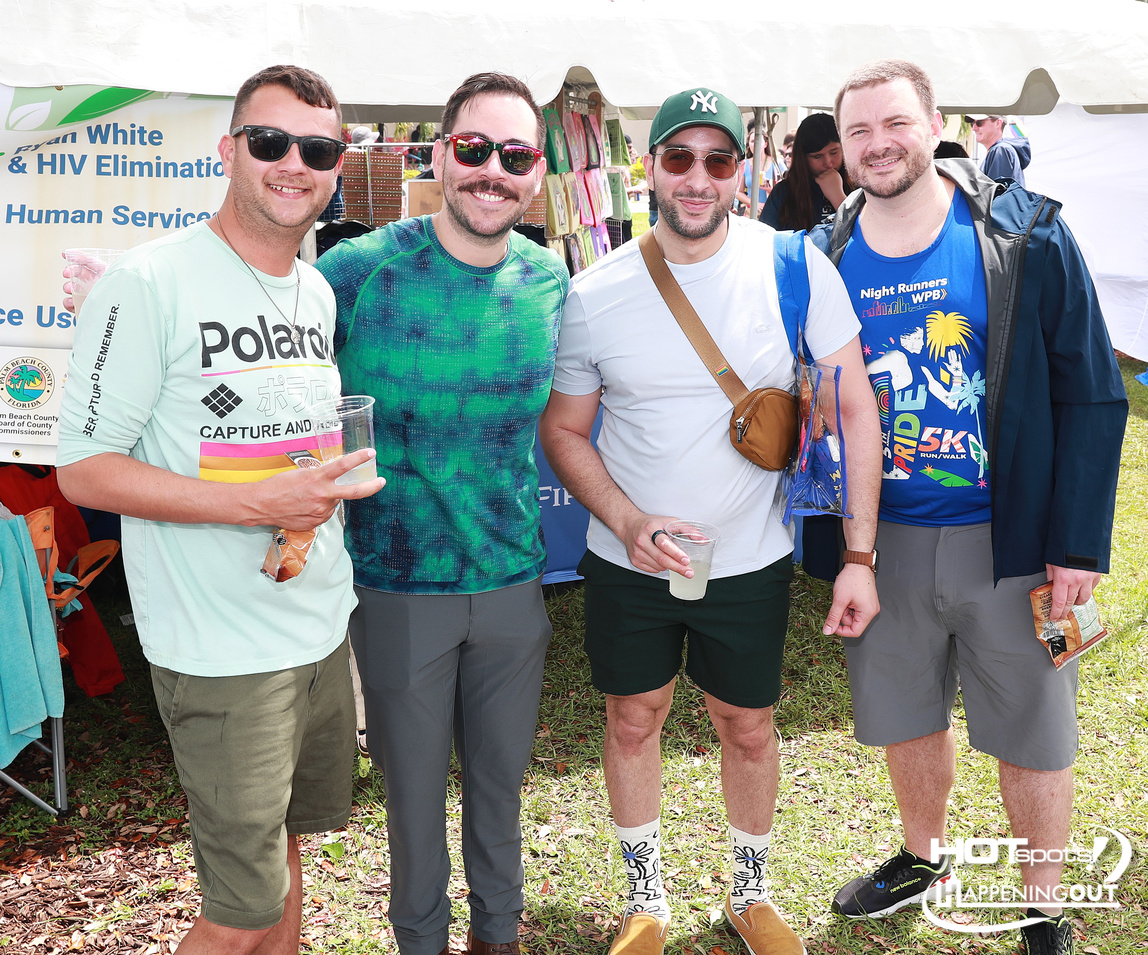 Four men posing together at a daytime outdoor health fair under a white tent, holding cups and smiling at the camera.