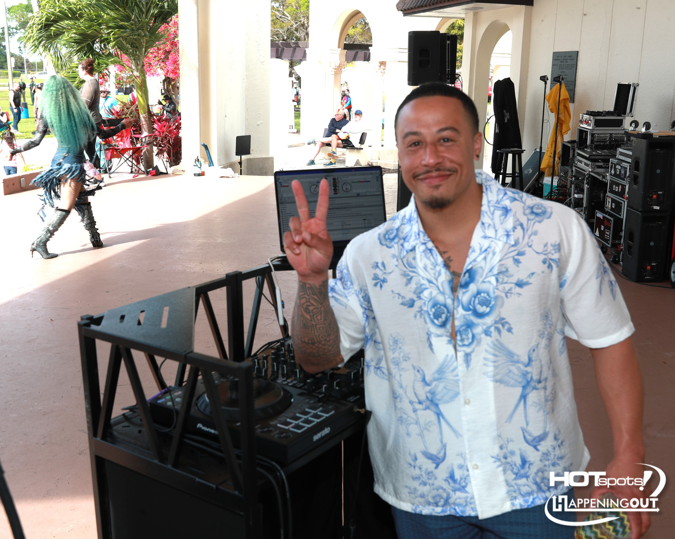 DJ booth setup at an outdoor event; man in a blue floral shirt shows a peace sign beside the mixing console, with dancers in the background.