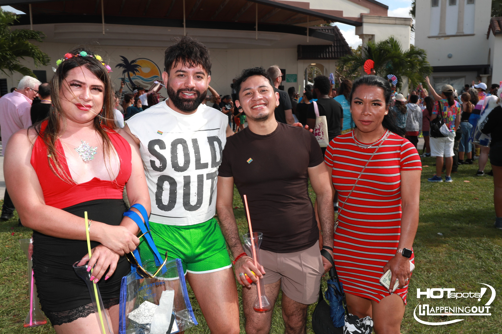 Four friends pose together at a sunny outdoor festival, each holding a drink and wearing bright outfits.
