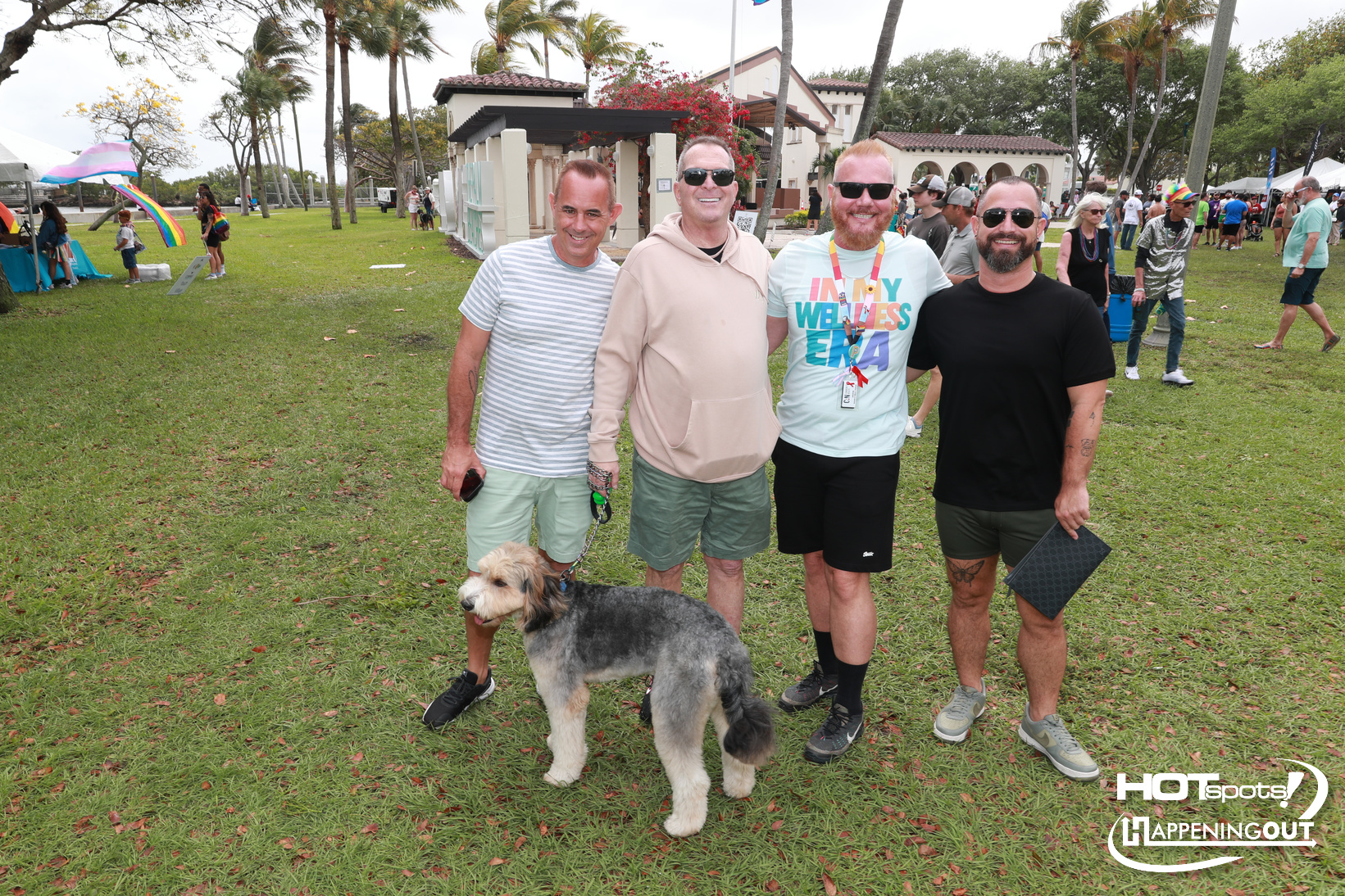 Four men posing with a gray dog at an outdoor park festival, smiling for the camera with tents and people in the background on a sunny day.