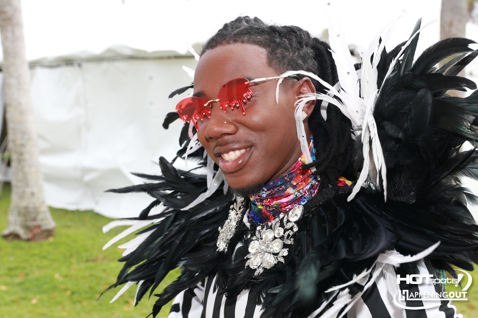 Smiling performer in a black-and-white feather costume with red heart-shaped sunglasses and sparkling jewelry at an outdoor event.
