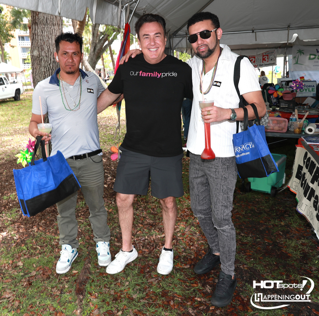 Three men pose together at an outdoor festival under a tent, smiling; center man wears a black shirt that says 'our family pride'.