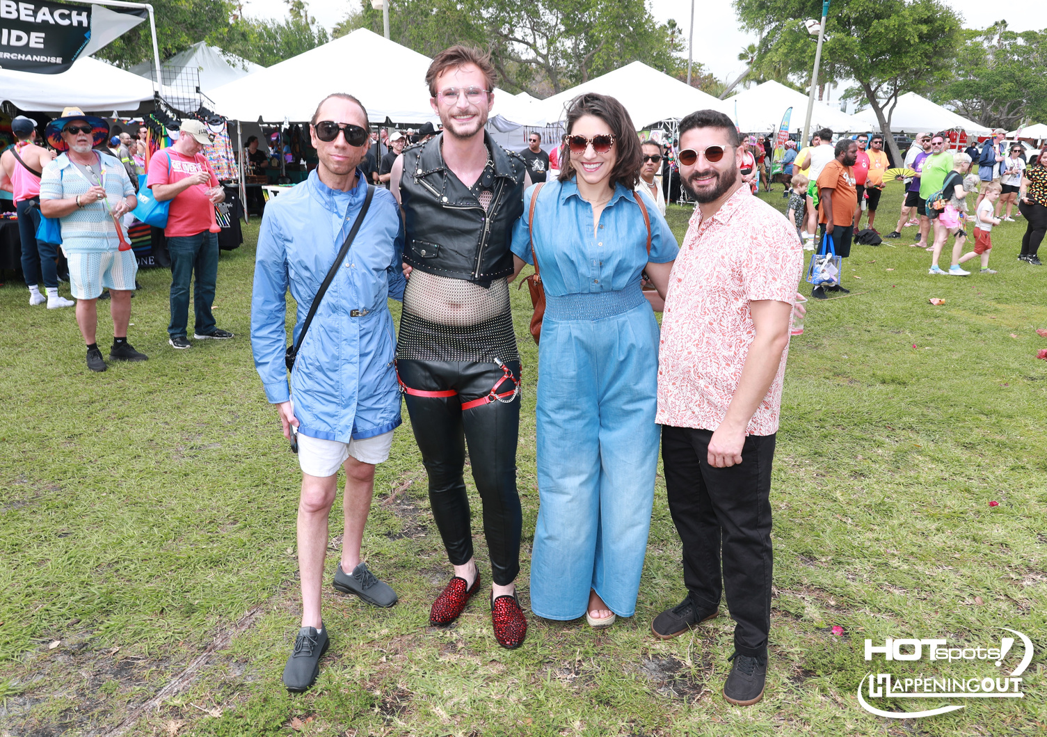 Four friends pose together on a sunny festival lawn, wearing bright, eclectic outfits with tents behind them.