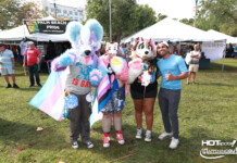 Group of four people in colorful fursuits posing at a Palm Beach Pride festival with vendor tents in the background, banner reading 'Palm Beach Pride'.