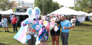 Group of four people in colorful fursuits posing at a Palm Beach Pride festival with vendor tents in the background, banner reading 'Palm Beach Pride'.