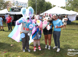 Group of four people in colorful fursuits posing at a Palm Beach Pride festival with vendor tents in the background, banner reading 'Palm Beach Pride'.