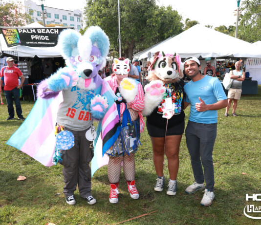 Group of four people in colorful fursuits posing at a Palm Beach Pride festival with vendor tents in the background, banner reading 'Palm Beach Pride'.