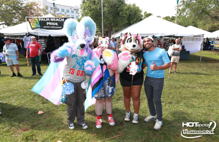 Group of four people in colorful fursuits posing at a Palm Beach Pride festival with vendor tents in the background, banner reading 'Palm Beach Pride'.