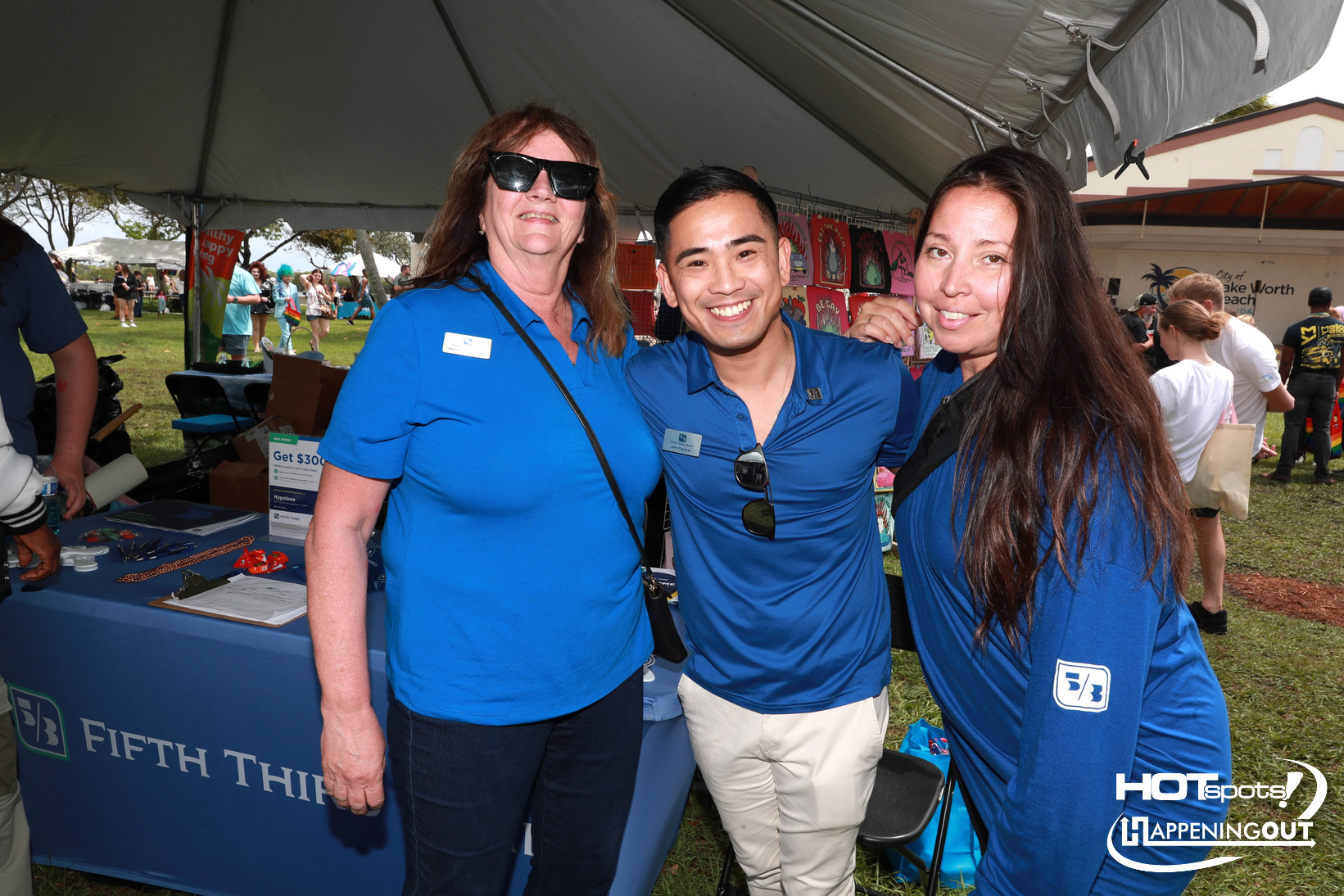 Three event staff in blue shirts pose together under a tent at an outdoor fair; table with brochures and a Fifth Third banner visible behind them.