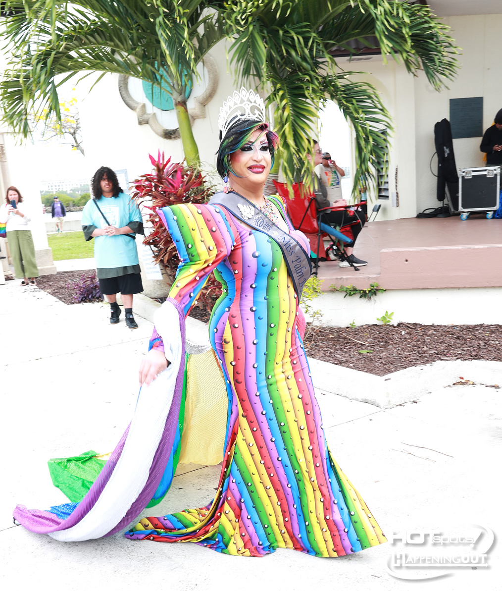 Drag queen in a long rainbow-striped gown with a silver crown and sash, posing outdoors at an event.