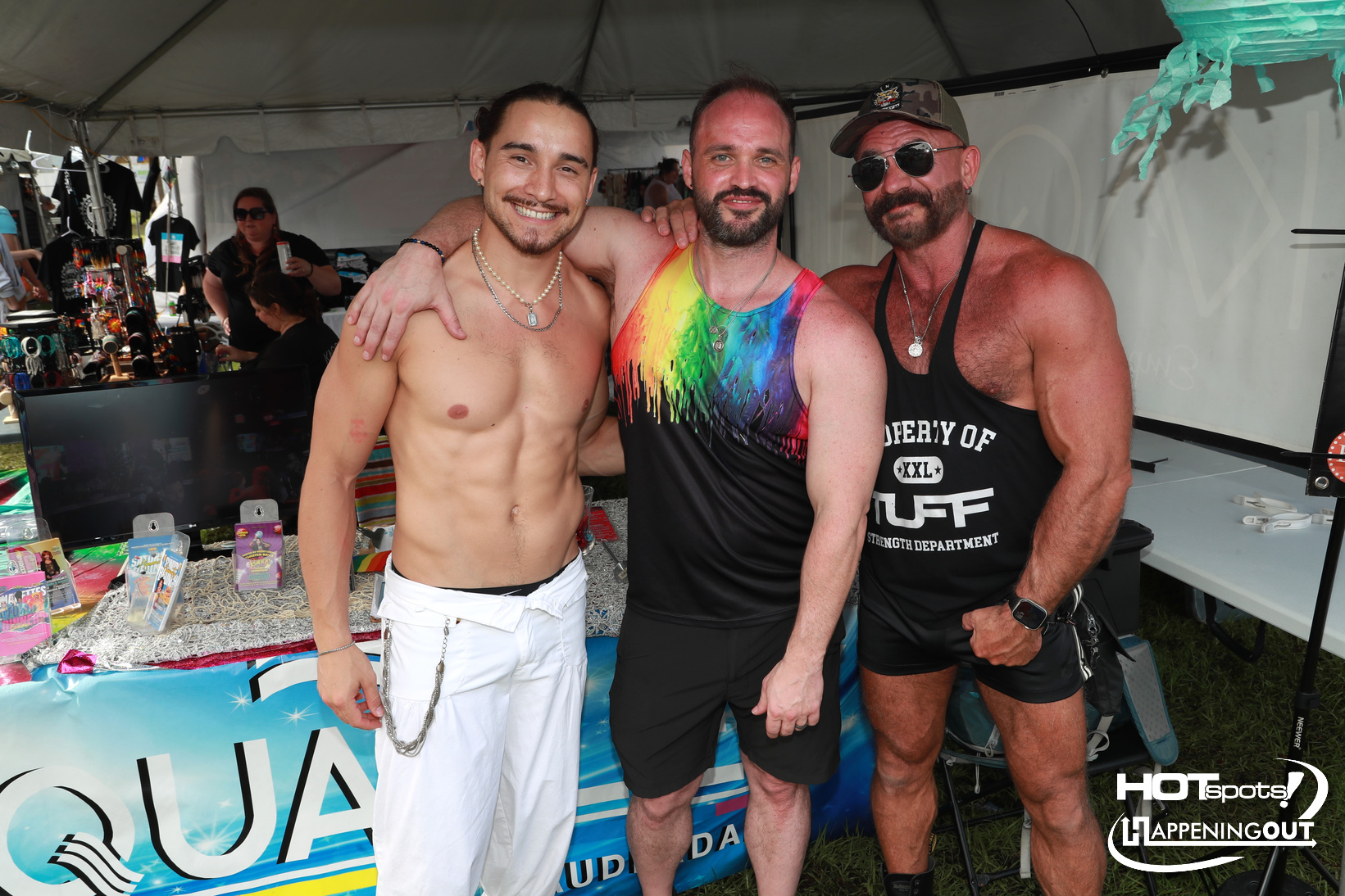 Three muscular men posing arm-in-arm at a festival/vendor booth, with rainbow shirt center and black tank right, smiles at camera.