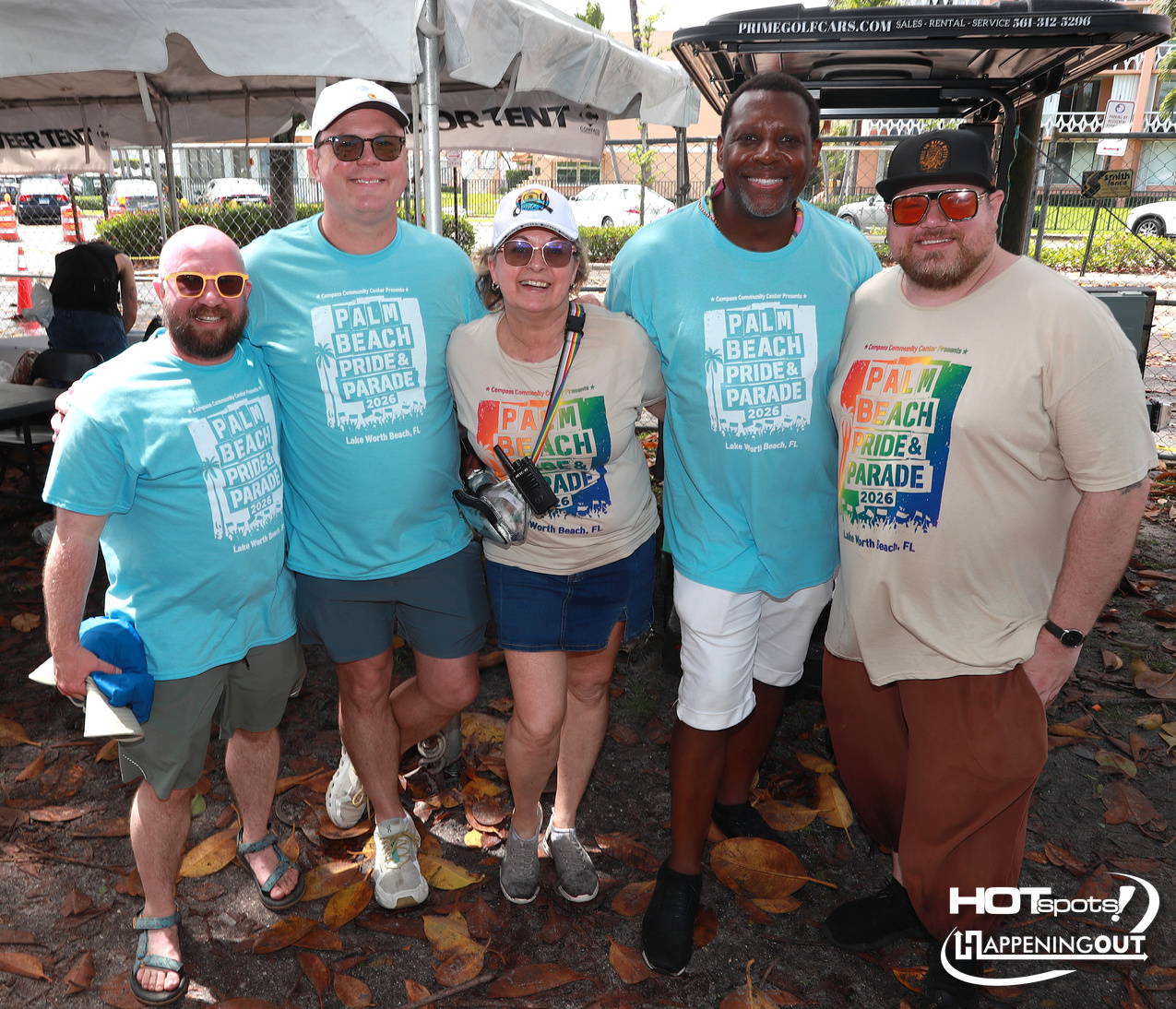 Group of five smiling adults posing together at an outdoor Palm Beach Pride & Parade event, with tents and autumn leaves in the background.