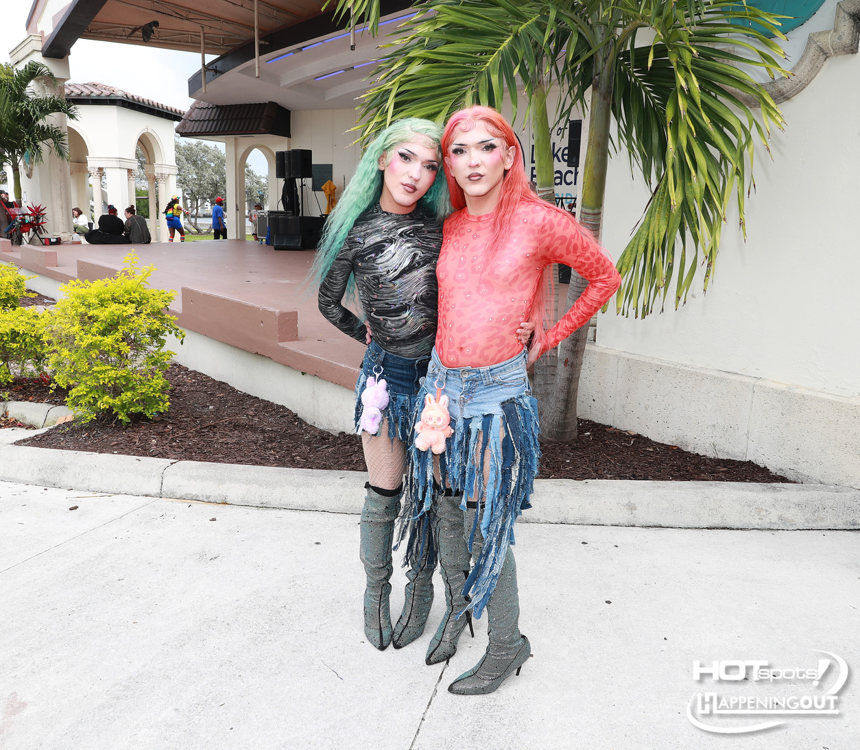 Two drag performers pose together outdoors in fringed denim skirts and knee-high boots, colorful hair—green and pink—standing near a stage and palm trees.