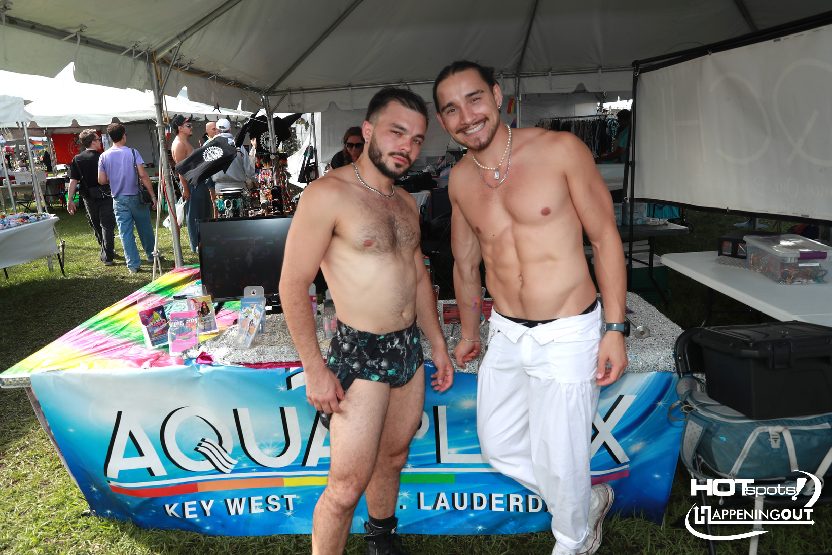 Two shirtless men pose and smile at an outdoor market booth with a bright Aqua West banner behind them.