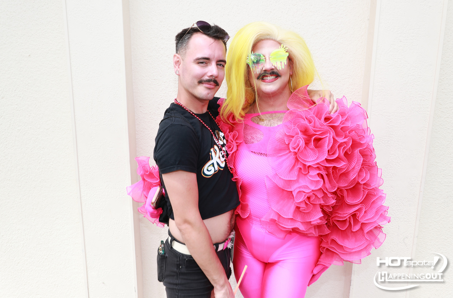 Two people pose together in colorful costumes: a man in a black shirt and beaded necklace standing beside a person in a bright pink ruffled outfit with a yellow wig and neon sunglasses.