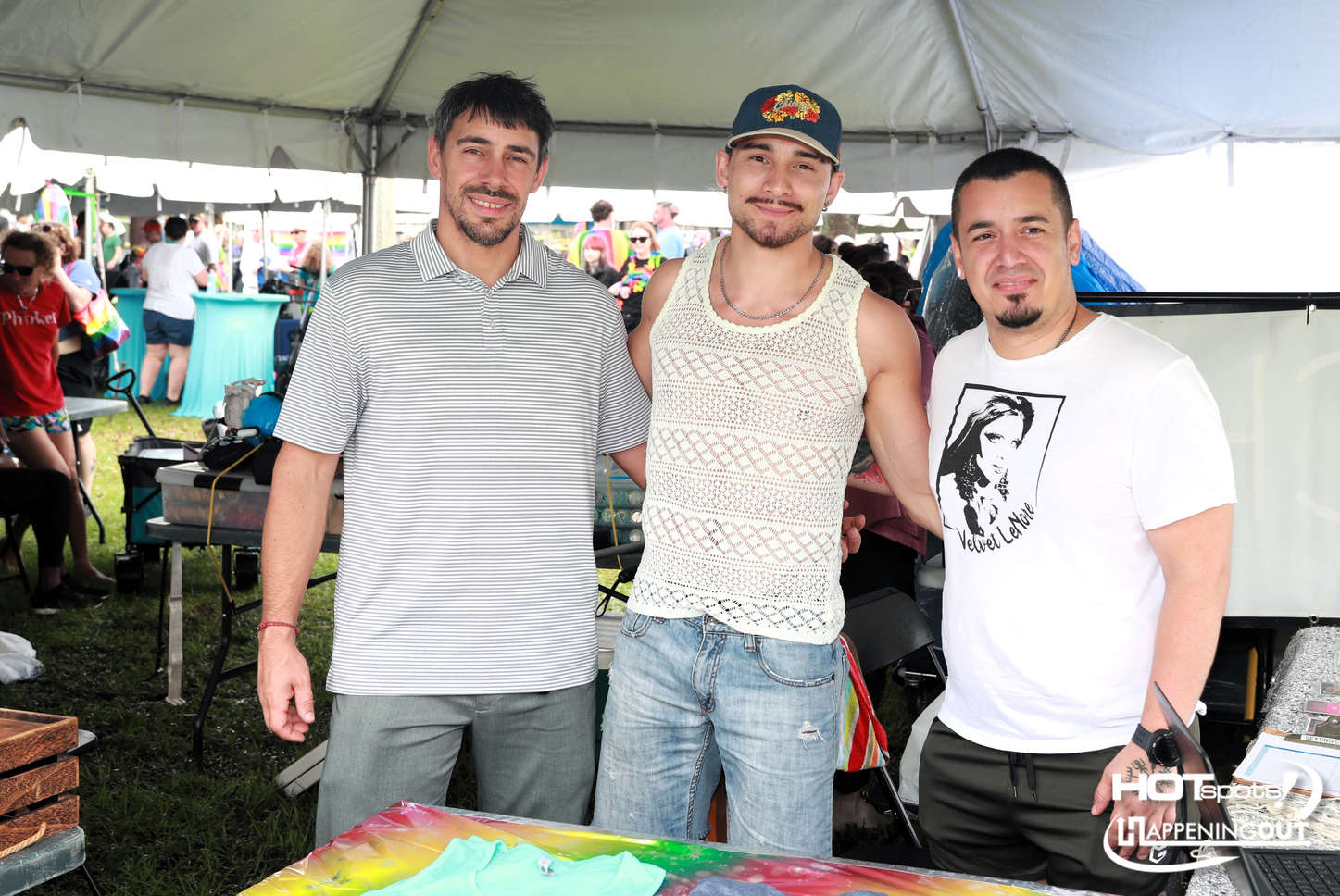 Three men pose for a photo at an outdoor festival booth under a canopy; left in a striped polo, center in a crochet tank top and cap, right in a white graphic tee.
