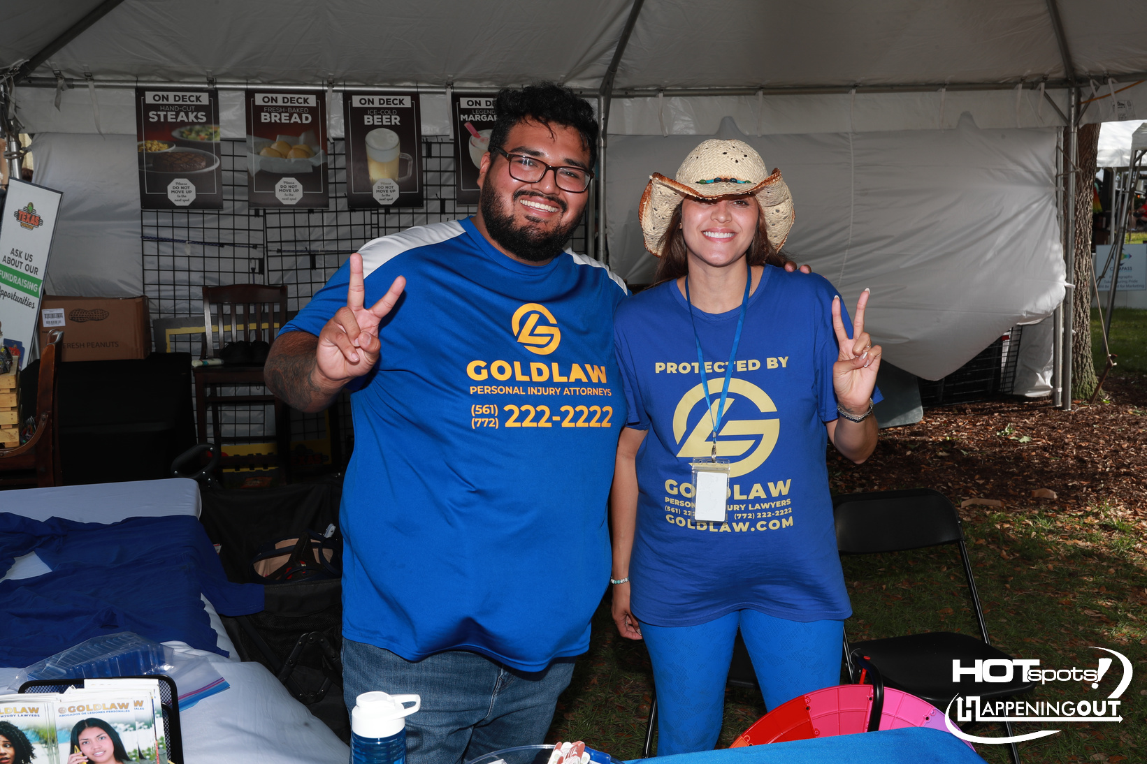 Two people in matching blue Goldlaw shirts smile and show peace signs at an outdoor event booth.