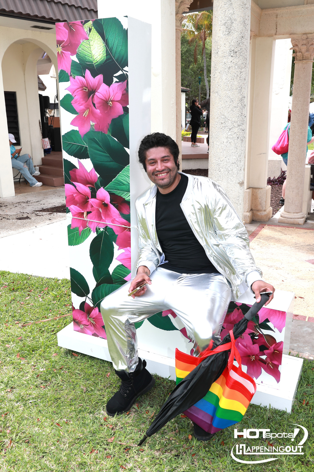 Smiling man in a metallic silver outfit sits beside a flower-patterned column at an outdoor event, holding a rainbow umbrella.