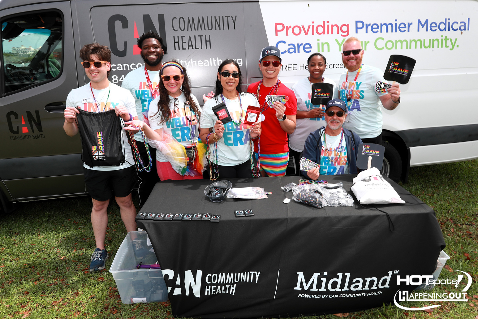 Group of volunteers at a CAN Community Health outreach tent, smiling and holding swag in front of a van with promotional banners.