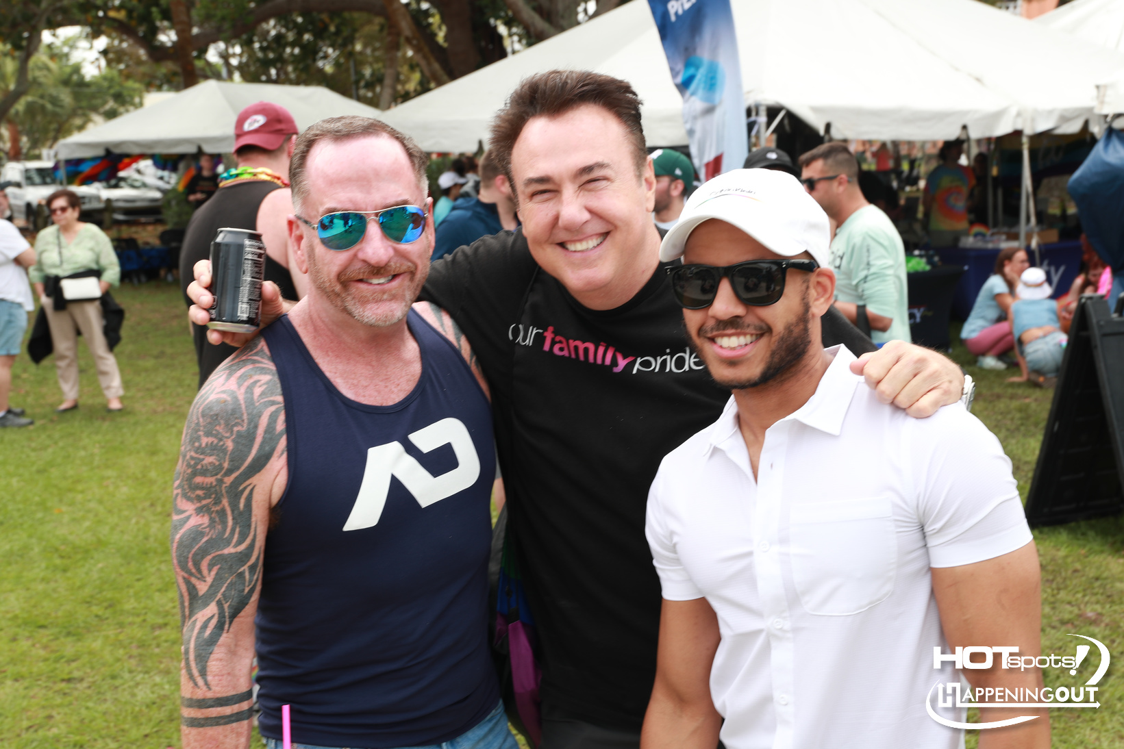 Three men smile for the camera at an outdoor festival, with tents and volunteers in the background. (
