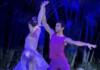 Two dancers hold hands overhead in a lift on stage, wearing purple outfits under blue lighting with tropical plants in the background, mid-performance.