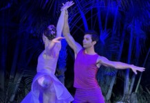Two dancers hold hands overhead in a lift on stage, wearing purple outfits under blue lighting with tropical plants in the background, mid-performance.