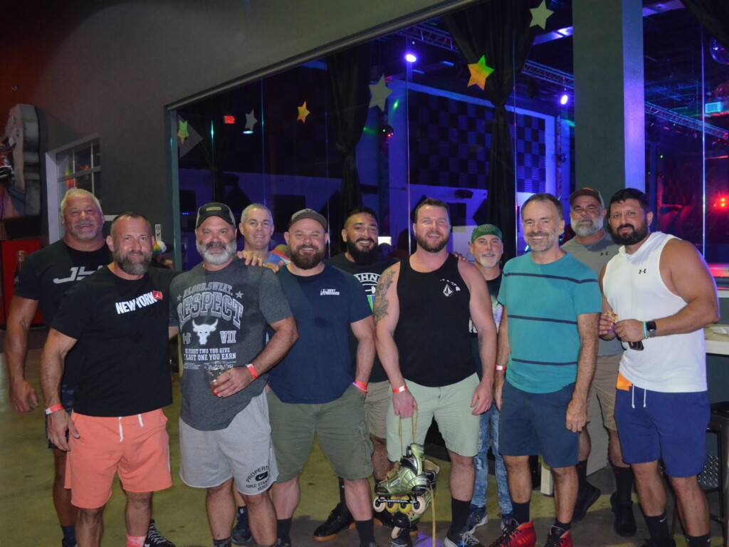 Group of men in casual athletic wear posing for a group photo at an indoor roller skating venue; roller skates visible on one person’s feet in front.