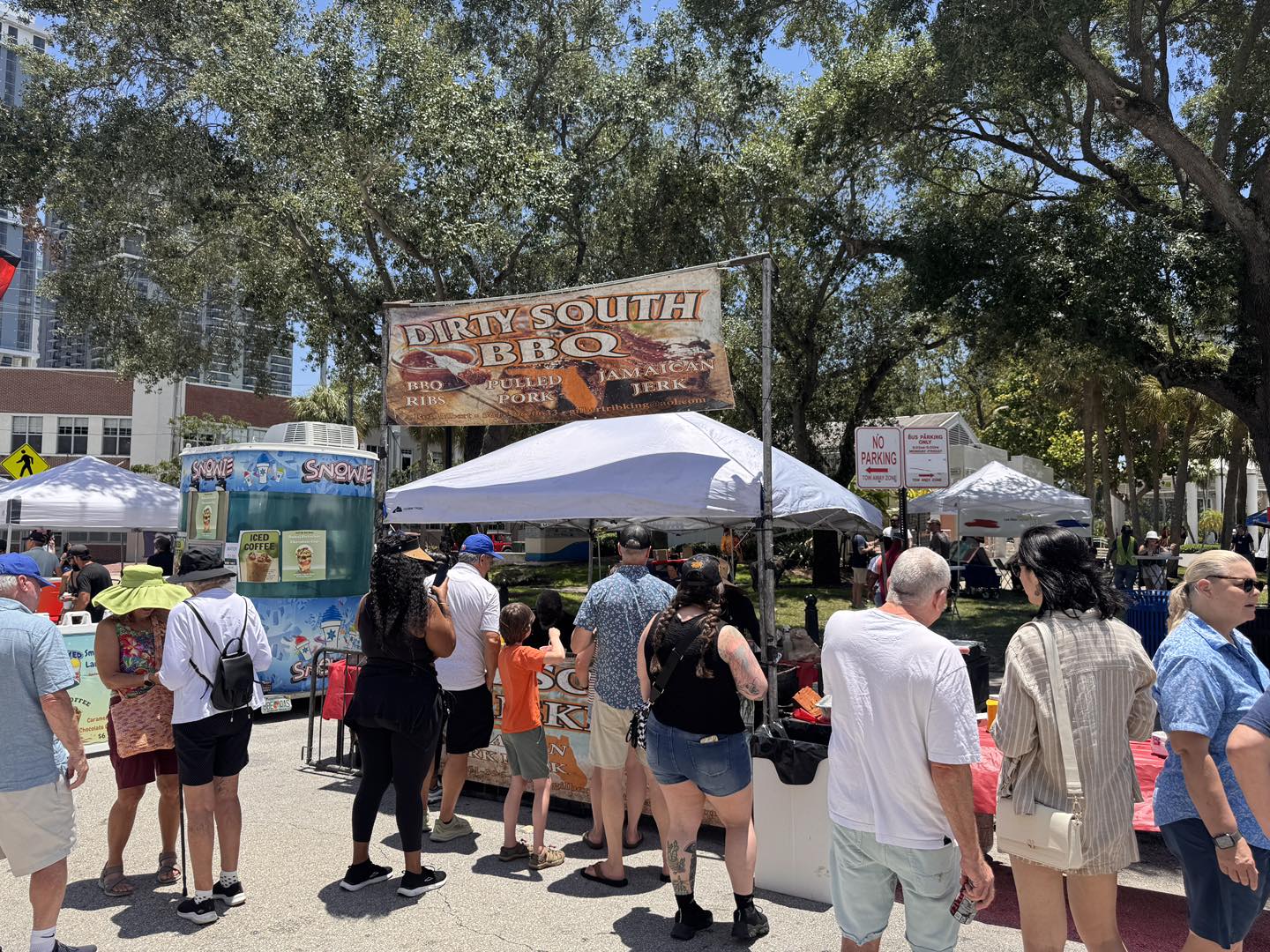 People line up at a Dirty South BBQ booth under a banner at an outdoor festival with tents and a snowie cart nearby.