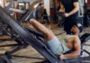 Man wearing a tank top and shorts uses a leg-press machine in a gym; trainer stands behind with a clipboard nearby.