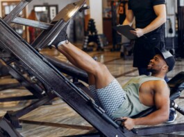 Man wearing a tank top and shorts uses a leg-press machine in a gym; trainer stands behind with a clipboard nearby.
