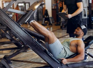Hot Bodies: Dynamic Training Man wearing a tank top and shorts uses a leg-press machine in a gym; trainer stands behind with a clipboard nearby.