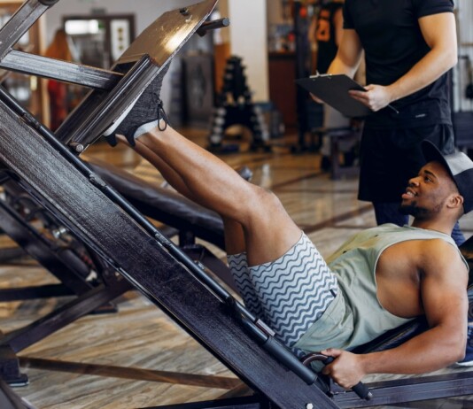 Man wearing a tank top and shorts uses a leg-press machine in a gym; trainer stands behind with a clipboard nearby.