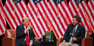 Two men in suits sit in armchairs on a stage with American flags behind them, each holding a microphone and chatting during a panel discussion.