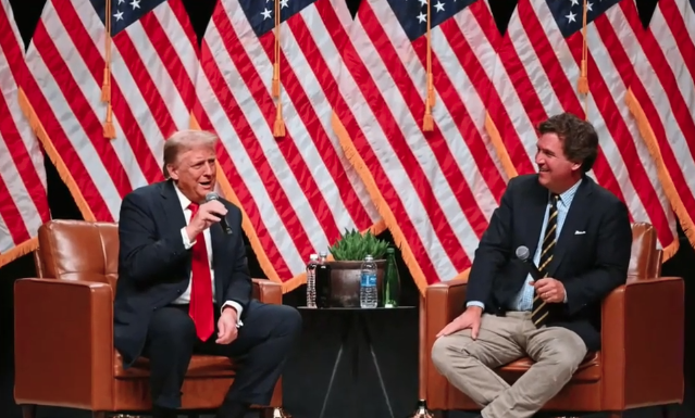 Tucker Carlson Tells Trump “You Have Failed,” Later Apologizes Two men in suits sit in armchairs on a stage with American flags behind them, each holding a microphone and chatting during a panel discussion.