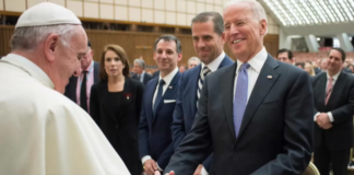 Religious leader in white robes shakes hands with a man in a dark suit at a formal event, with several attendees in business attire in the background.