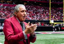 Man in a red suit speaks with his hands clapped together on a football field, packed stadium in the background.