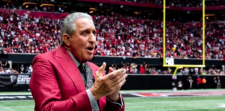 Man in a red suit speaks with his hands clapped together on a football field, packed stadium in the background.