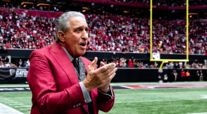 Man in a red suit speaks with his hands clapped together on a football field, packed stadium in the background.