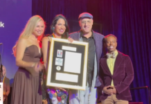 Group of four people on stage posing with a framed award plaque, smiling for the camera; purple backdrop and sign reading 'Greater Wilton Manors' visible.